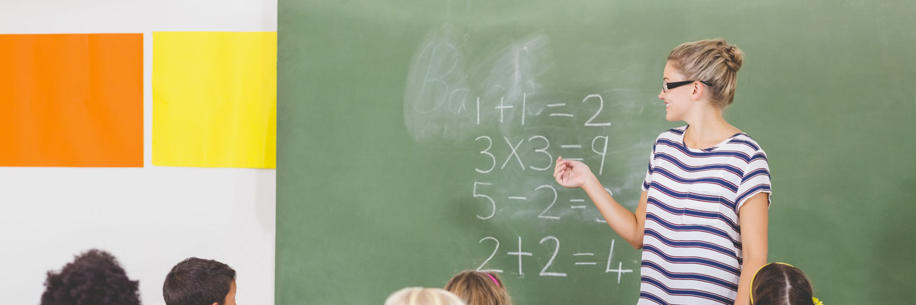 teacher pointing to chalkboard in front of class full of students